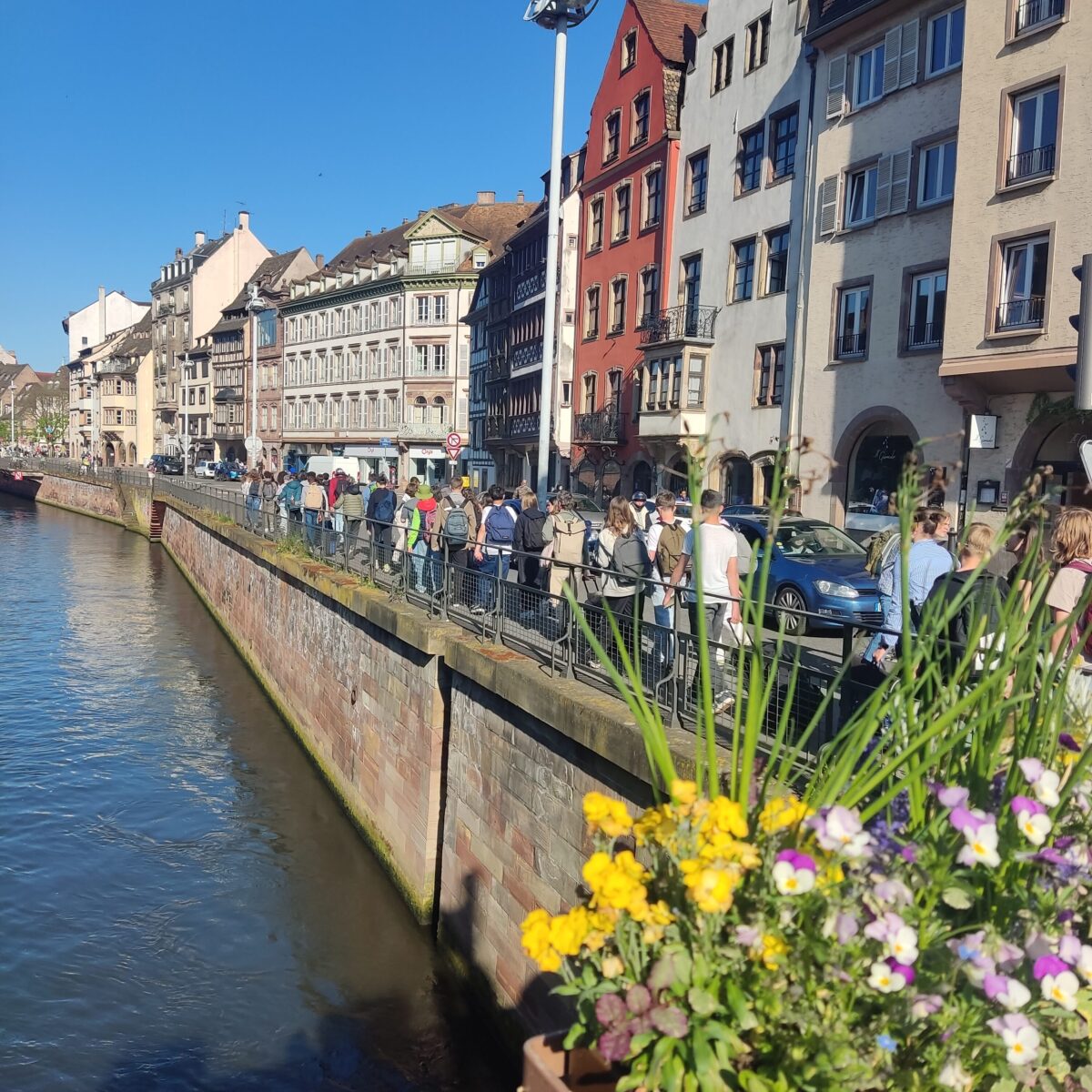 Eine belebte Straße mit Personen entlang eines Flusses, bunten Blumen im Vordergrund, sonniger Tag.