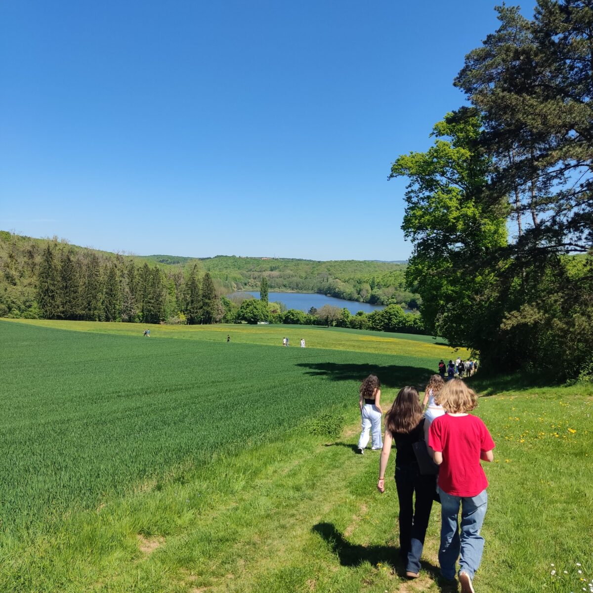 Personen spazieren auf einem Pfad durch grüne Landschaft, mit blauem Himmel und einem See im Hintergrund.