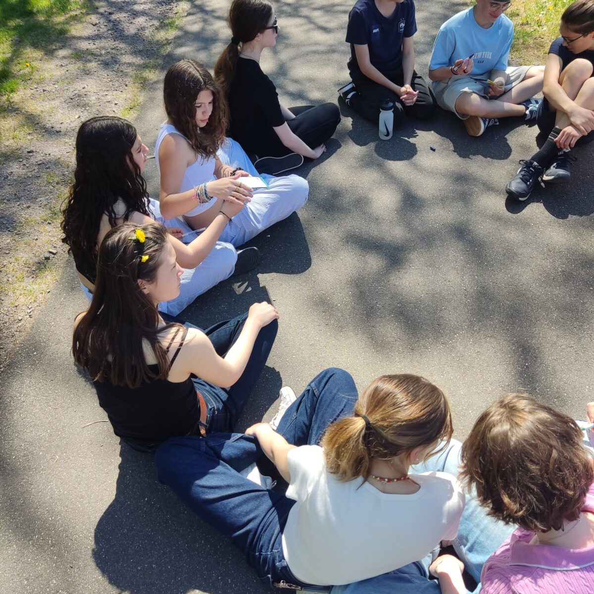 Eine Gruppe junger Menschen sitzt im Kreis auf einem Weg im Freien und lacht gemeinsam.