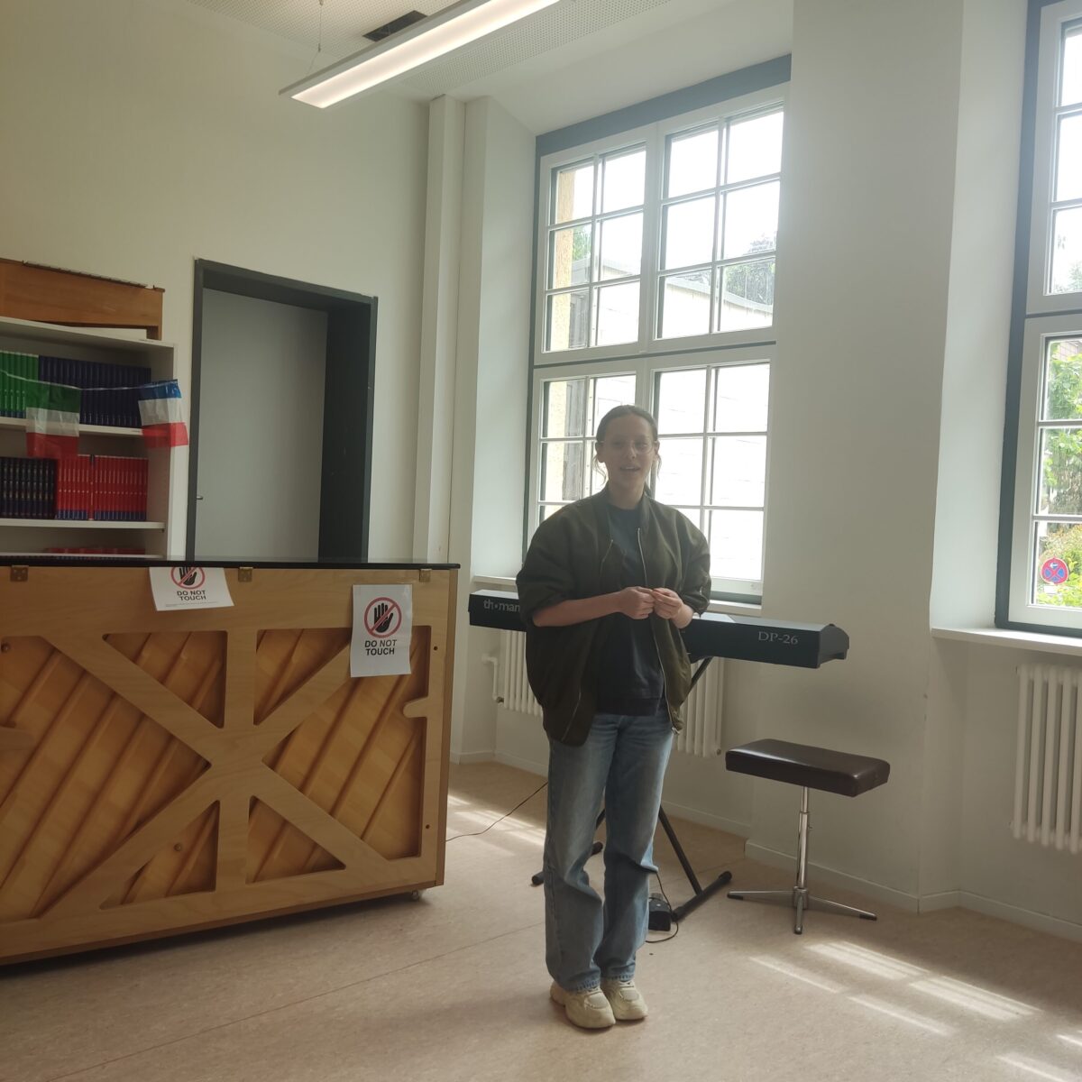 Eine Person lächelt in einem Raum mit Klavier und bunten Büchern, trägt eine grüne Jacke.