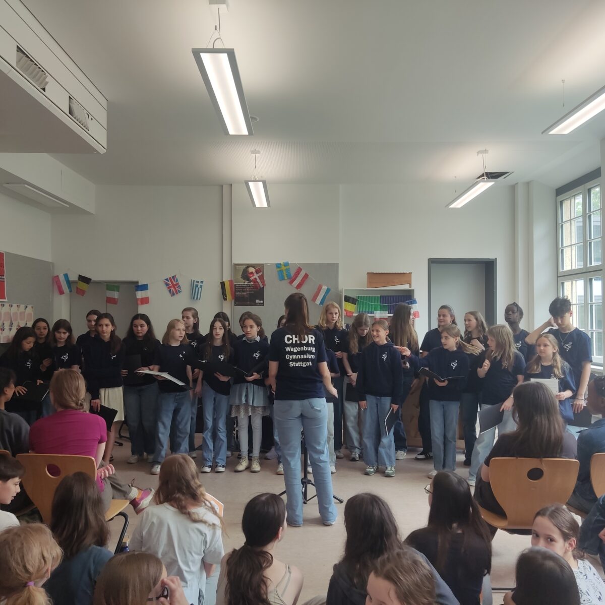 Eine Gruppe Jugendlicher im Chor, dirigiert von einer Leiterin in Jeans und schwarzem T-Shirt.