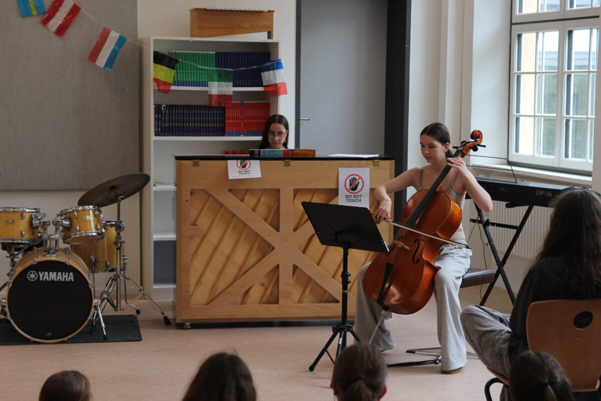 Eine junge Frau spielt Cello, eine andere sitzt am Klavier; im Hintergrund sind bunte Flaggen.