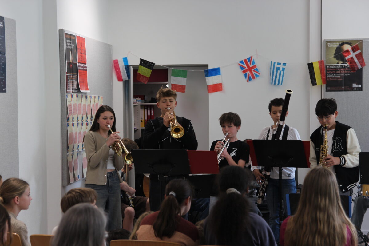 Sechs junge Musiker, konzentriert, mit verschiedenen Blasinstrumenten, europäische Flaggen im Hintergrund.