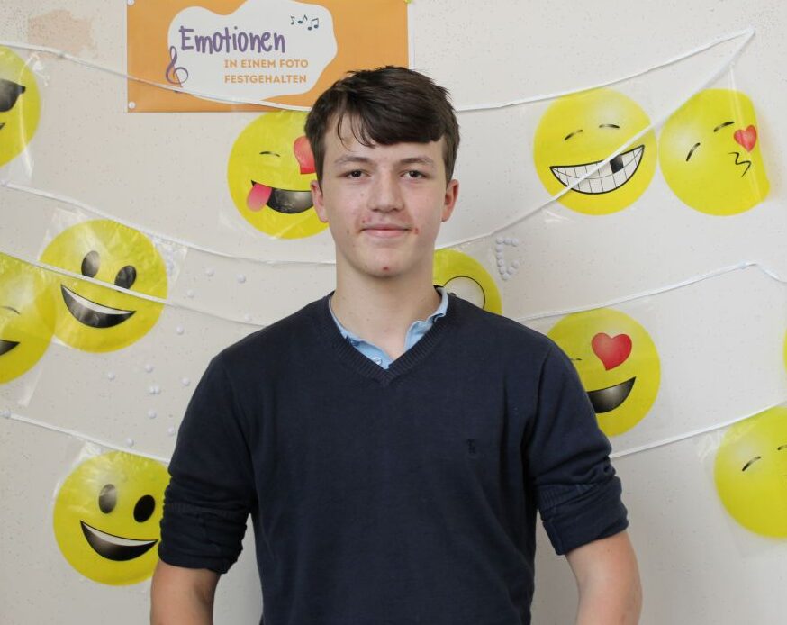 Ein junger Mann lächelt vor einem Hintergrund mit gelben Emojis und einem Schild "Emotionen".