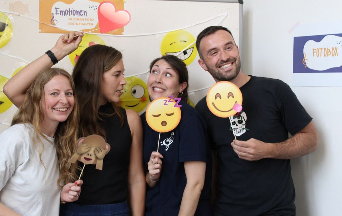Drei Frauen und ein Mann lächeln und halten Emoji-Requisiten vor einer weißen Wand mit Smiley-Stickern.