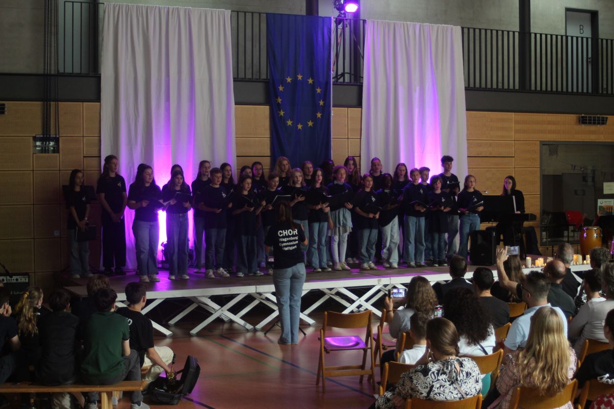 Gruppenauftritt auf Bühne, Personen in schwarzer Kleidung, EU-Flagge im Hintergrund, Publikum sitzt.