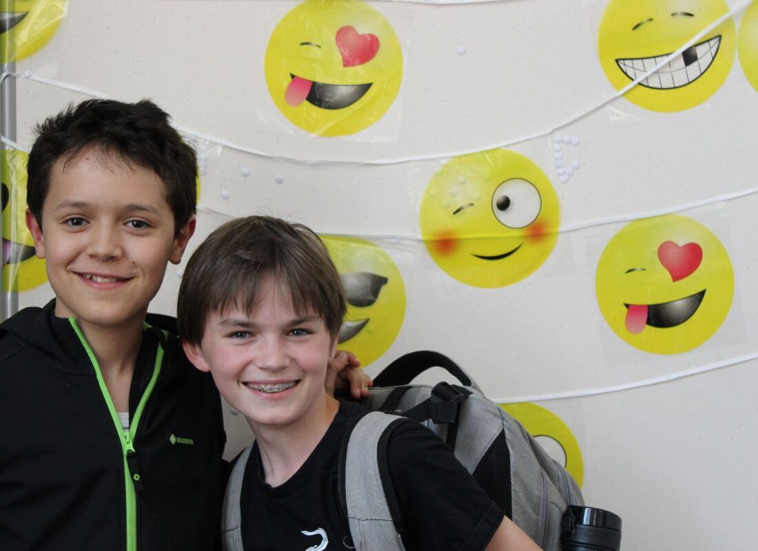 Zwei lächelnde Jungen vor einer Emoji-Wand, einer mit Rucksack, die Emojis auf weißem Hintergrund.