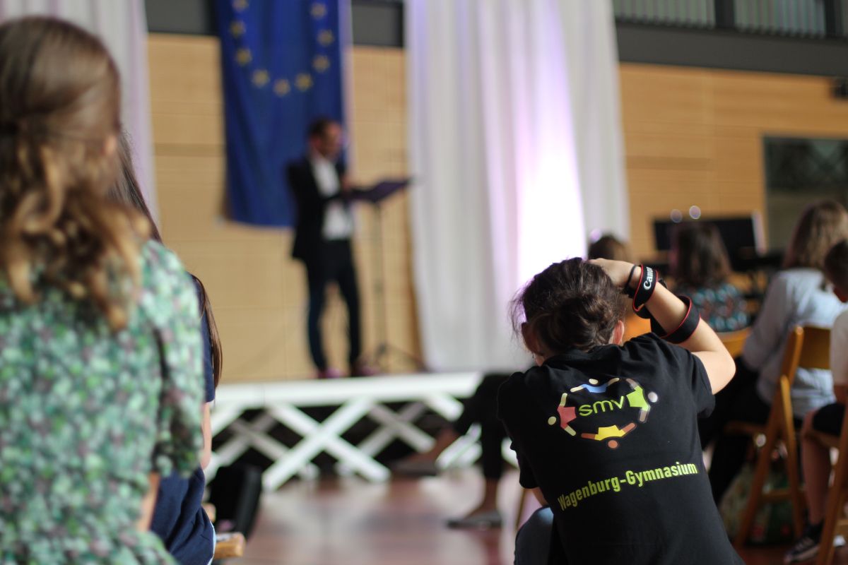 Eine Frau hört einem Redner mit EU-Flagge zu, trägt ein schwarz-grünes T-Shirt.
