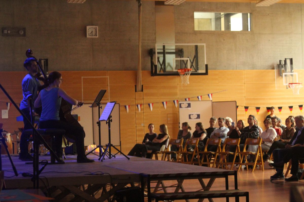 Eine Cellistin und ein Cellist spielen auf einer Bühne vor Publikum in einer Turnhalle.