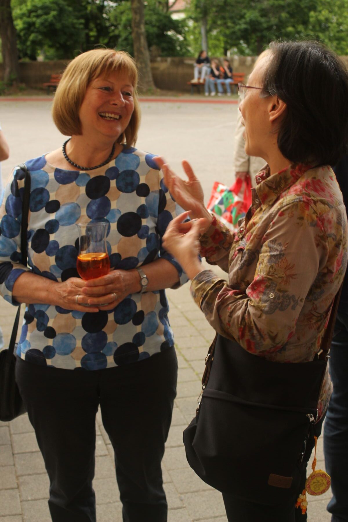 Zwei lächelnde Frauen, stehend, eine im bunten Polka-Dot-Shirt mit Getränk, die andere in Blumenbluse.
