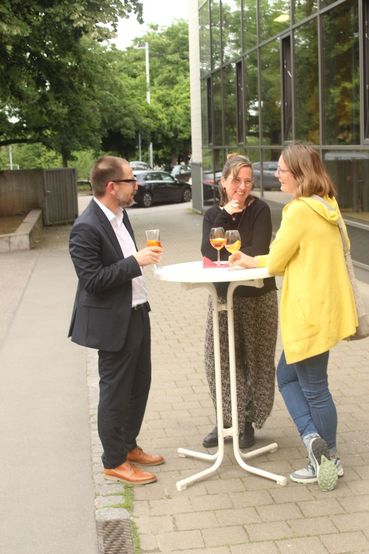 Drei Personen stehen lächelnd an einem Tisch im Freien mit Getränken, eine trägt einen gelben Mantel.