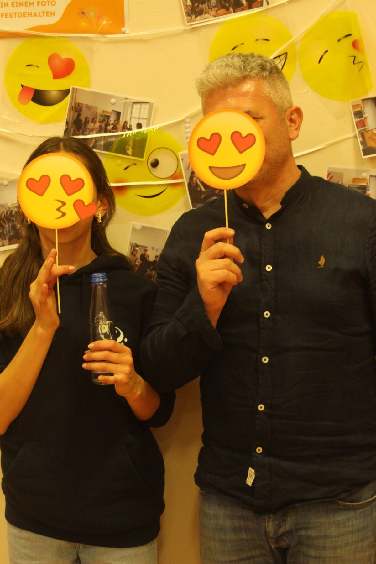 Zwei Personen halten Emoji-Wandmasken mit Herzaugen vor ihr Gesicht, im Hintergrund gelbe Emojis.