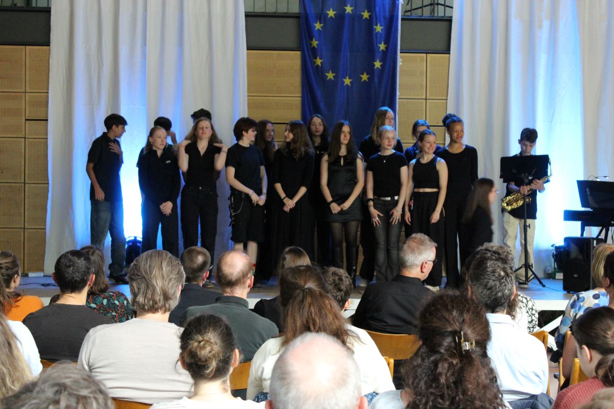 Eine Gruppe junger Personen in Schwarz steht lächelnd auf einer Bühne vor einer EU-Flagge.