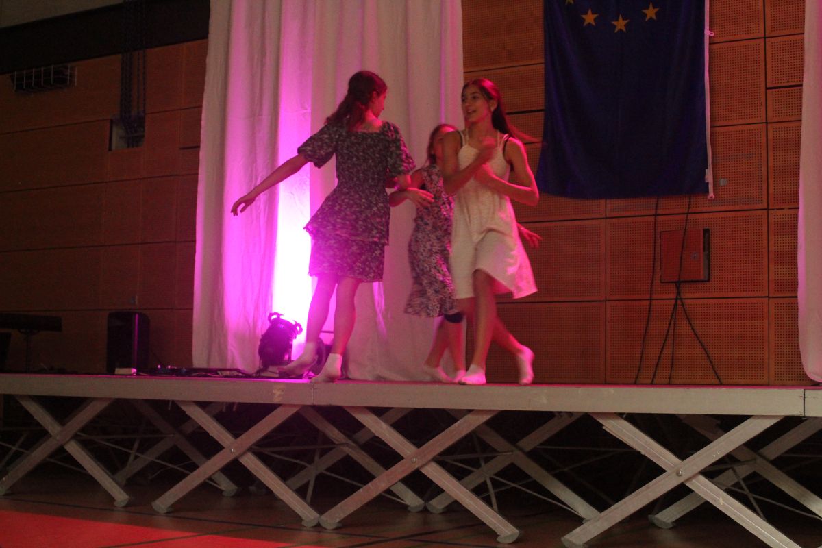 Drei Frauen tanzen fröhlich auf einer Bühne mit rosa Licht und EU-Flagge im Hintergrund.