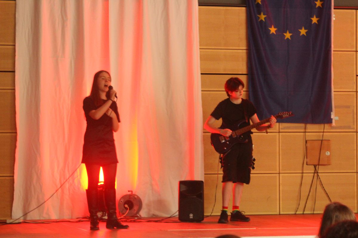 Eine Frau singt und ein Junge spielt Gitarre auf der Bühne, beide in schwarzer Kleidung.