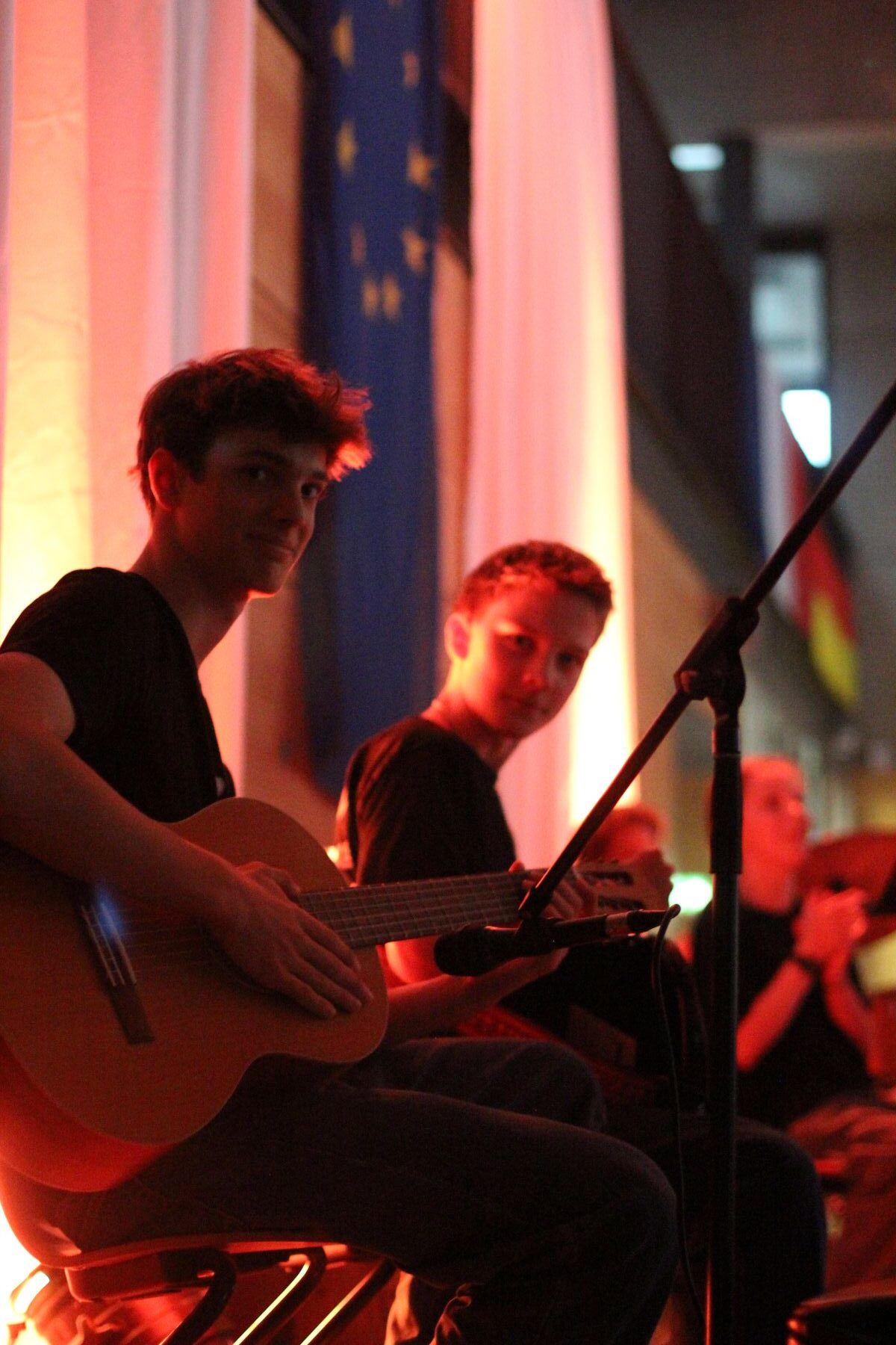 Zwei Männer spielen Gitarre und lächeln im warmen Licht auf einer Bühne mit EU-Flagge im Hintergrund.