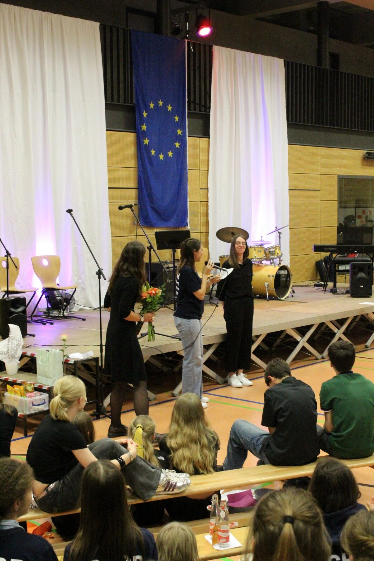 Eine Gruppe von Personen auf einer Bühne, eine Frau lächelt und hält Blumen, EU-Flagge im Hintergrund.