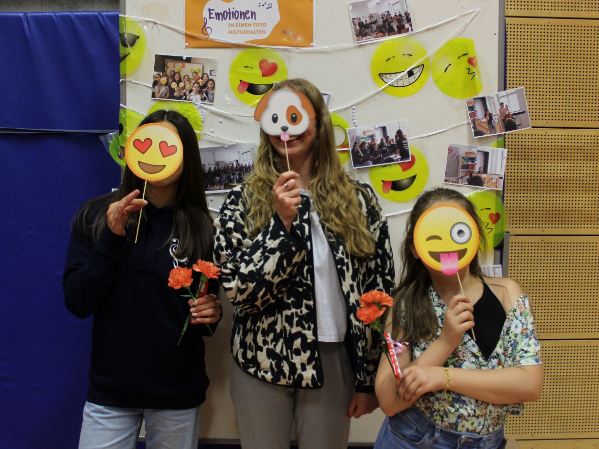 Drei Personen mit Emoji-Masken lächeln und halten Blumen vor einer bunten Emoji-Wand.
