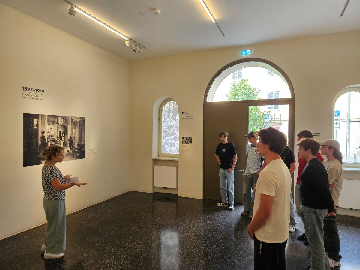 Eine Frau erklärt einer Gruppe junger Männer ein Foto in einem hellen Ausstellungsraum.