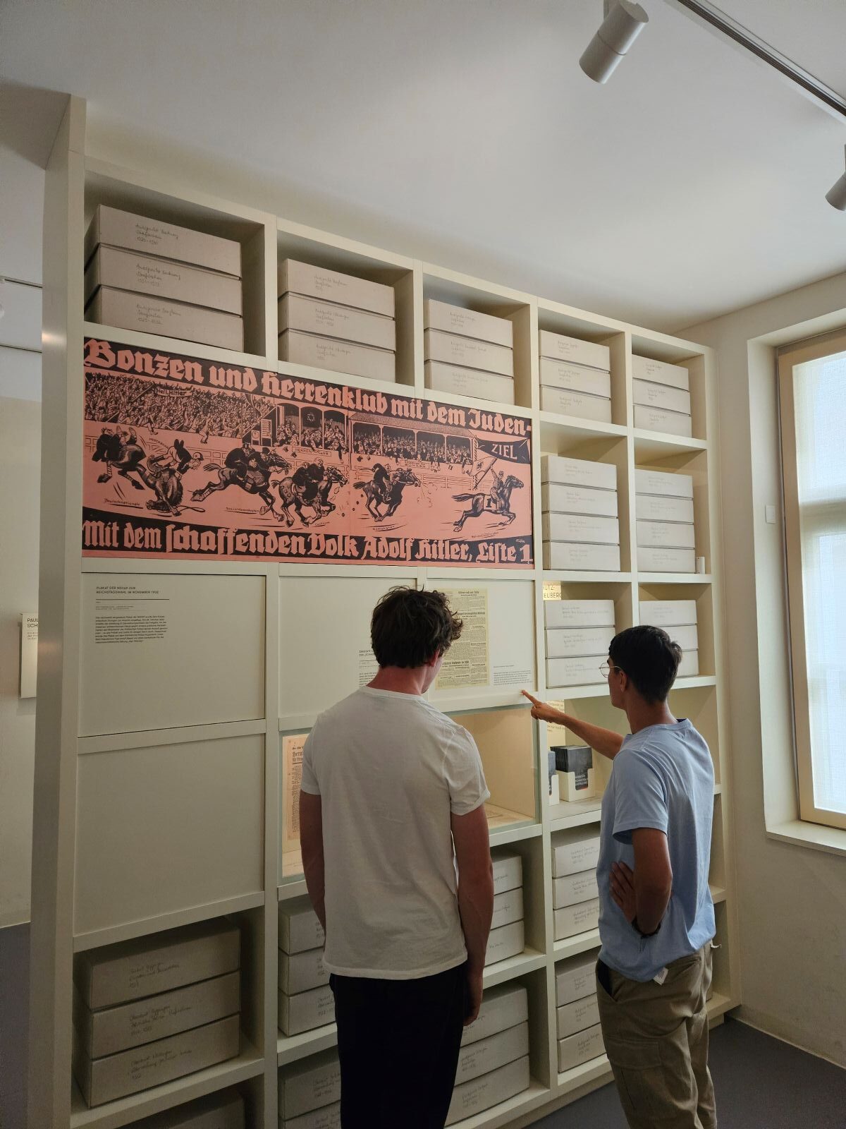 Zwei Männer im Museum betrachten ein Regal mit Kisten und ein historisches Plakat an der Wand.