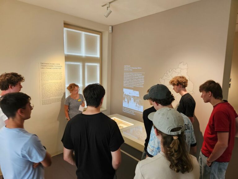 Gruppe junger Menschen in Museumsausstellung, interessiert, Frau erklärt Exponat. Hell erleuchteter Raum.
