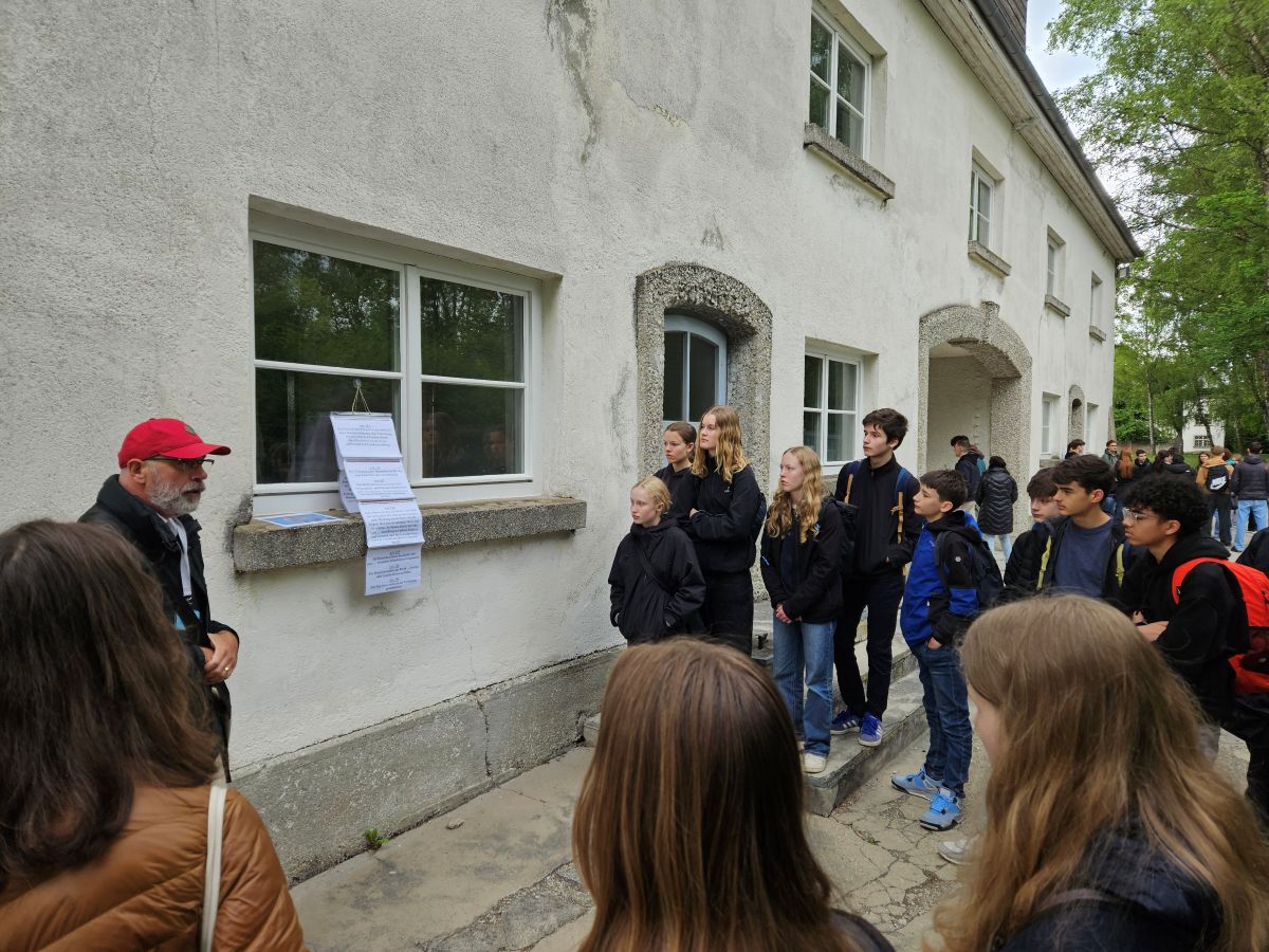 Älterer Mann erklärt Gruppe von Jugendlichen vor weißem Gebäude, trägt rote Mütze und schwarzen Mantel.