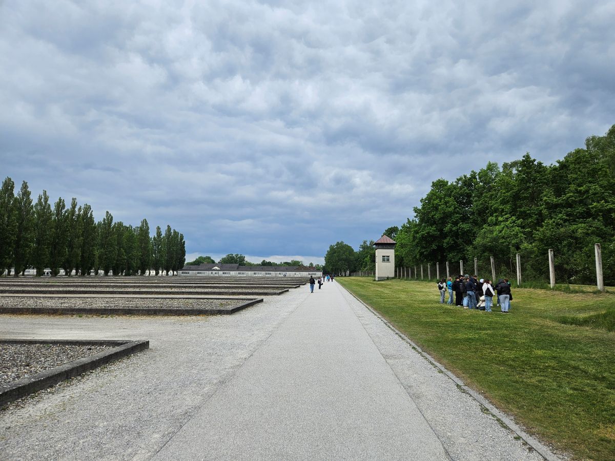 Weitläufige Gedenkstätte mit Besuchern, Wolkenhimmel, grüne Bäume, Kiesflächen und Pfade.