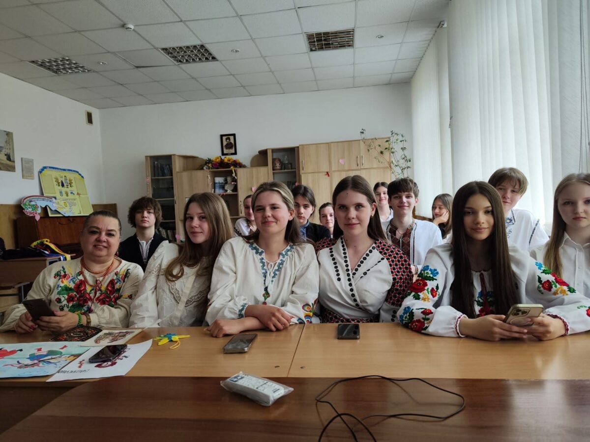 Eine Gruppe lächelnder Menschen in bestickter traditioneller Kleidung sitzt an Tischen in einem Klassenraum.