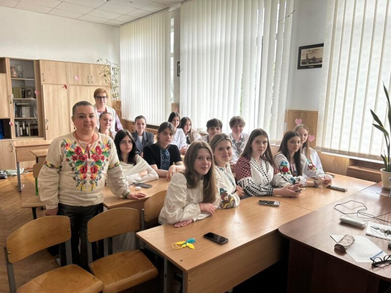 Eine Gruppe junger Menschen und eine ältere Frau lächeln sitzend in einem Klassenzimmer.