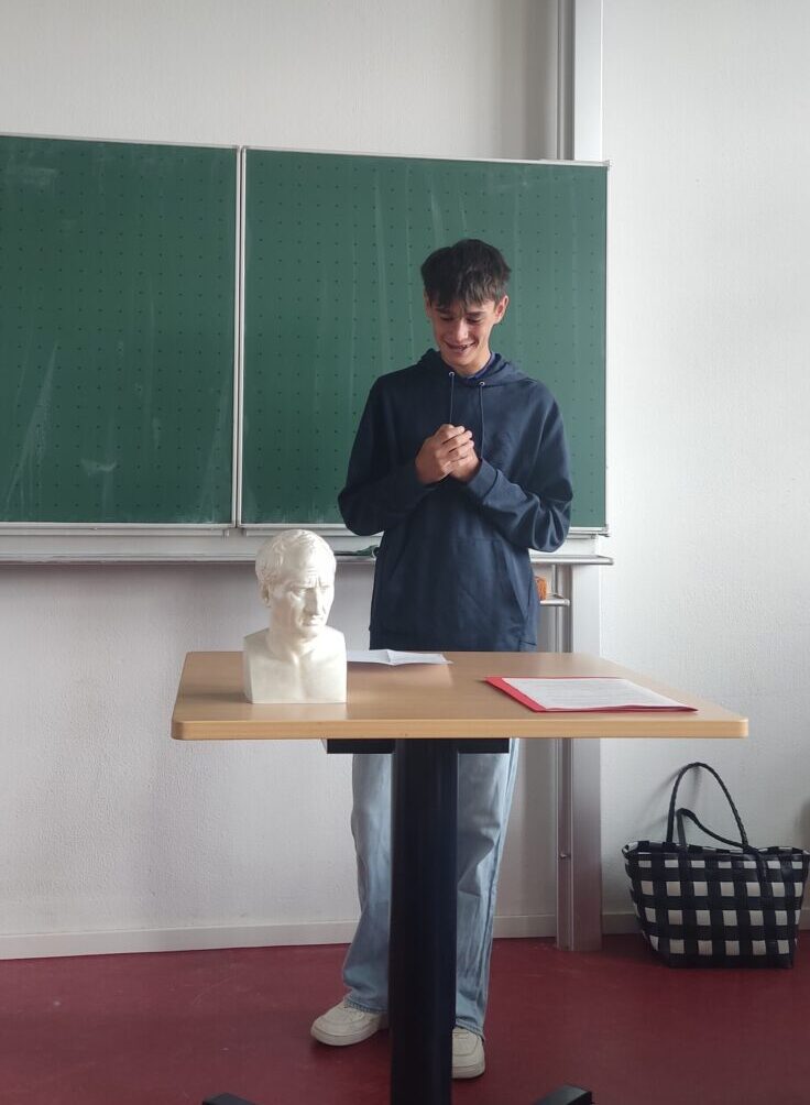Ein junger Mann lacht vor einer Tafel neben einer Büste und einem Tisch.