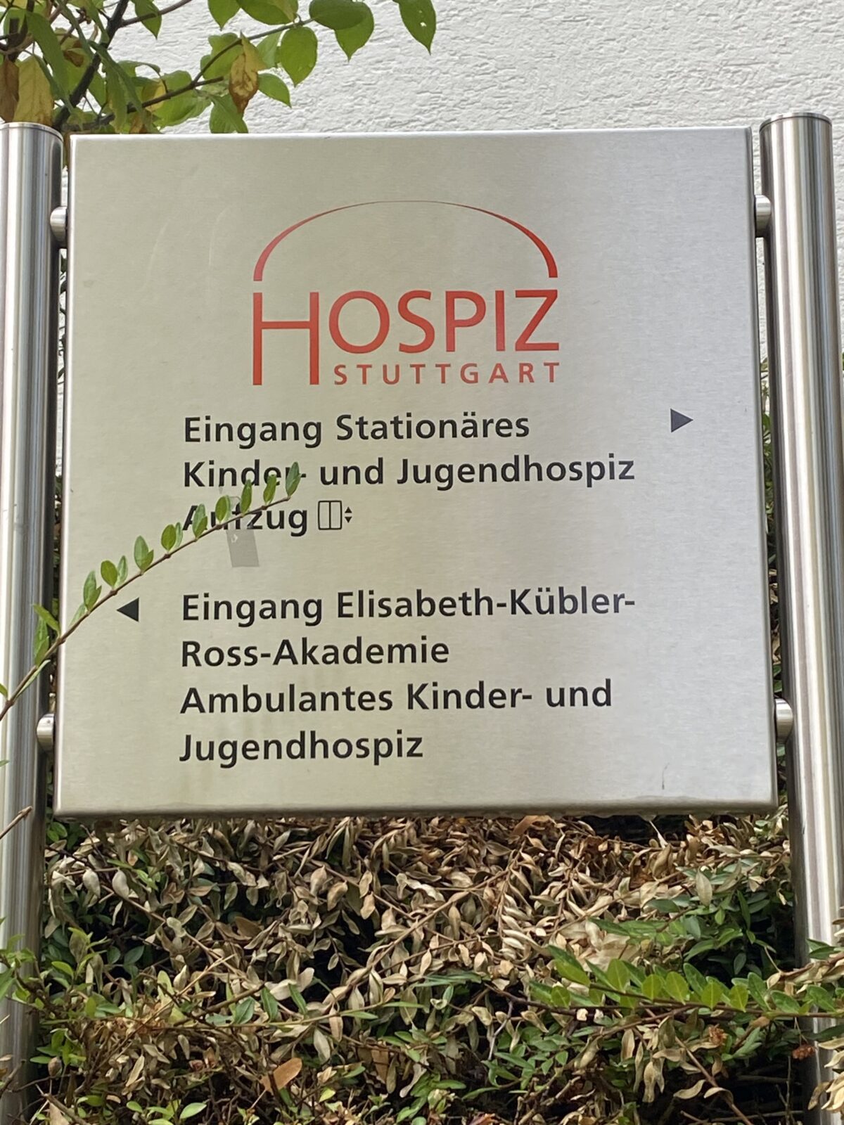 Schild des Hospiz Stuttgart in einem Garten mit Pflanzen im Vordergrund.