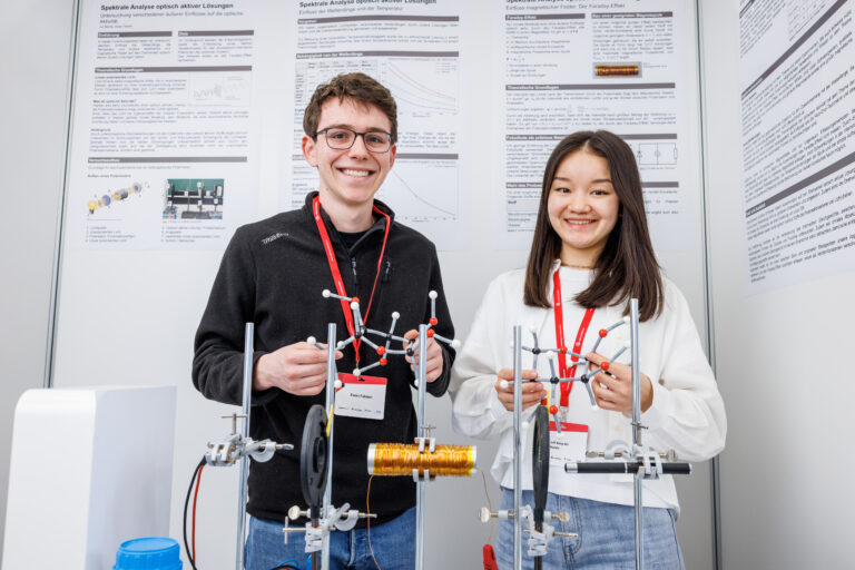 Zwei junge Erwachsene lächeln und präsentieren wissenschaftliche Modelle vor Postern.