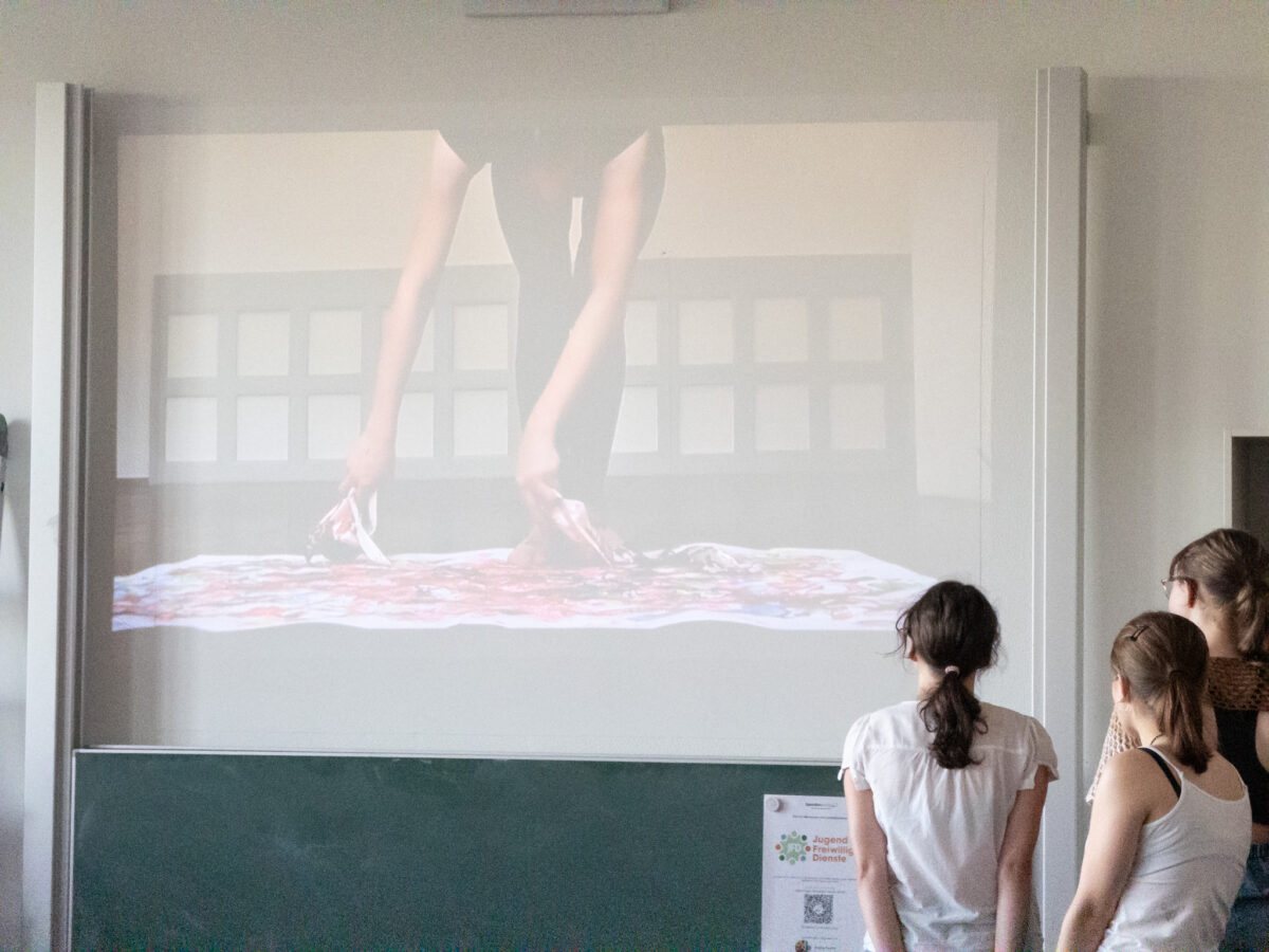 Drei Frauen betrachten ein Video, das Hände zeigt. Interessantes Kunstwerk auf großem Bildschirm.