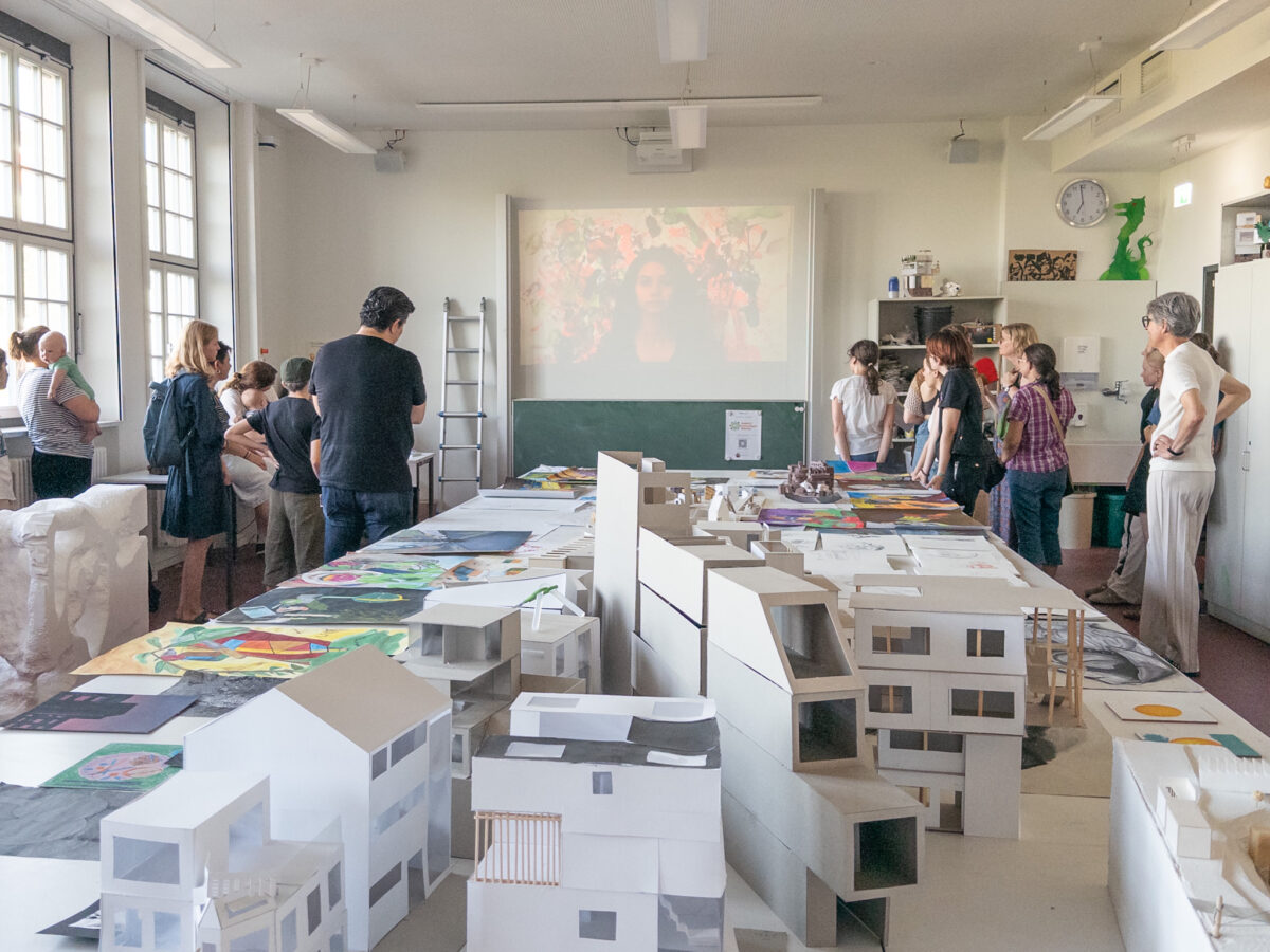 Menschen betrachten Kunst in einem hellen Raum mit Architekturmodellen und Gemälden.