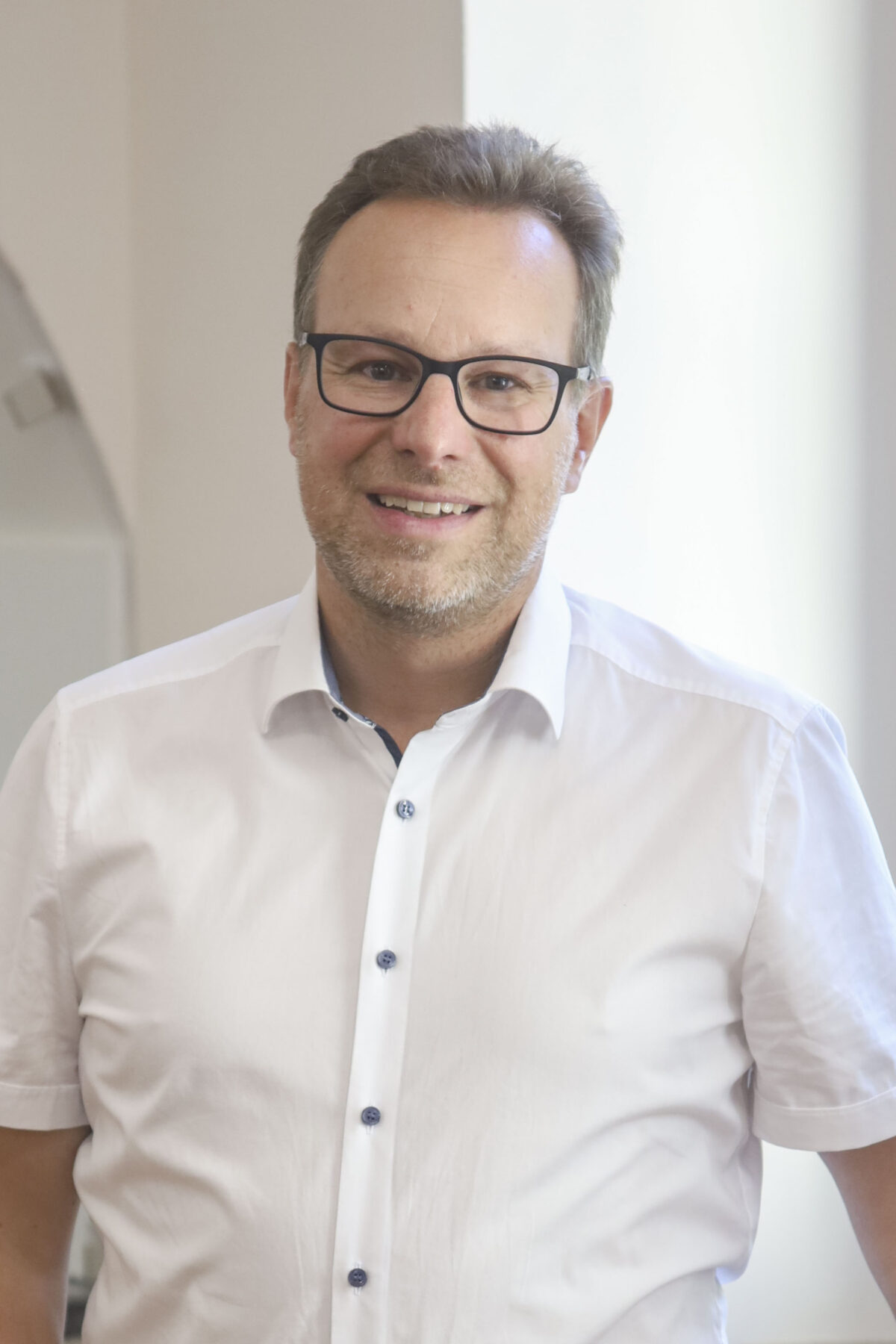 Ein Herr mit Brille lächelt, trägt ein weißes Hemd in einem hellen Raum.