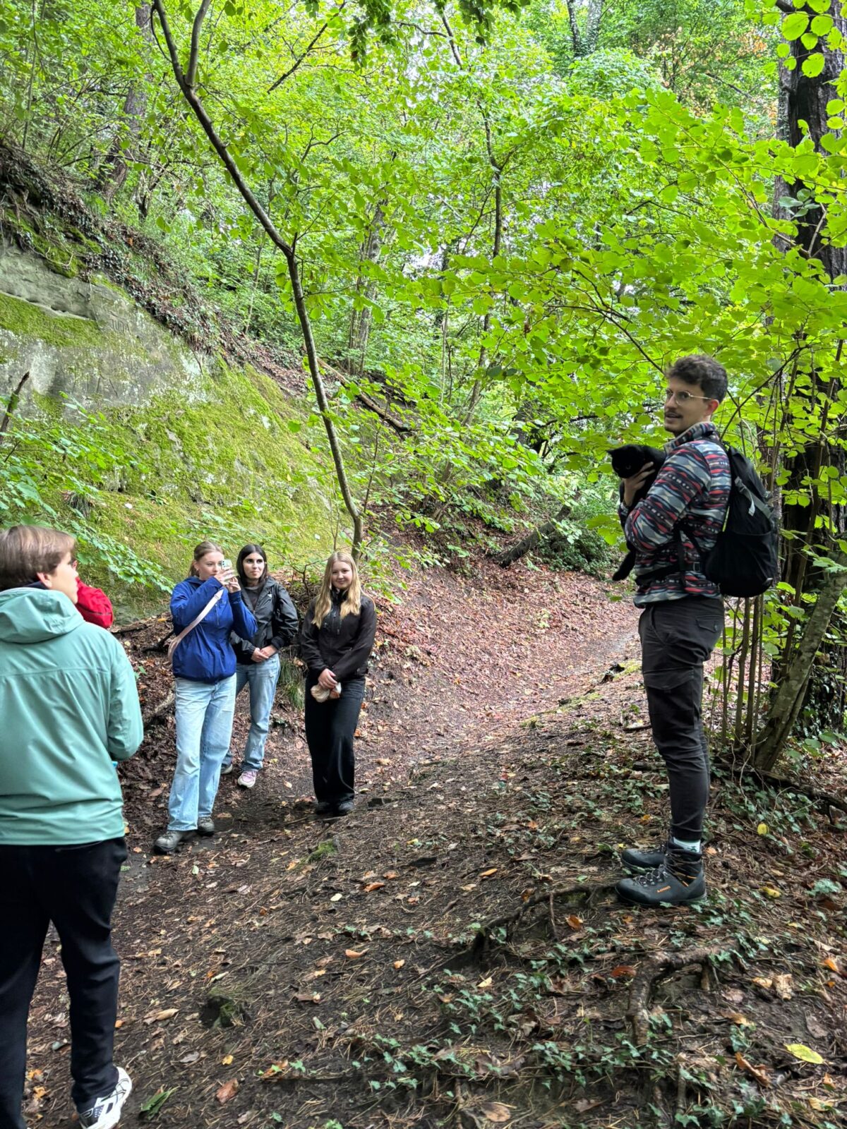 Eine Gruppe junger Menschen steht aufmerksam im grünen Wald, ein Mann hält einen Hund