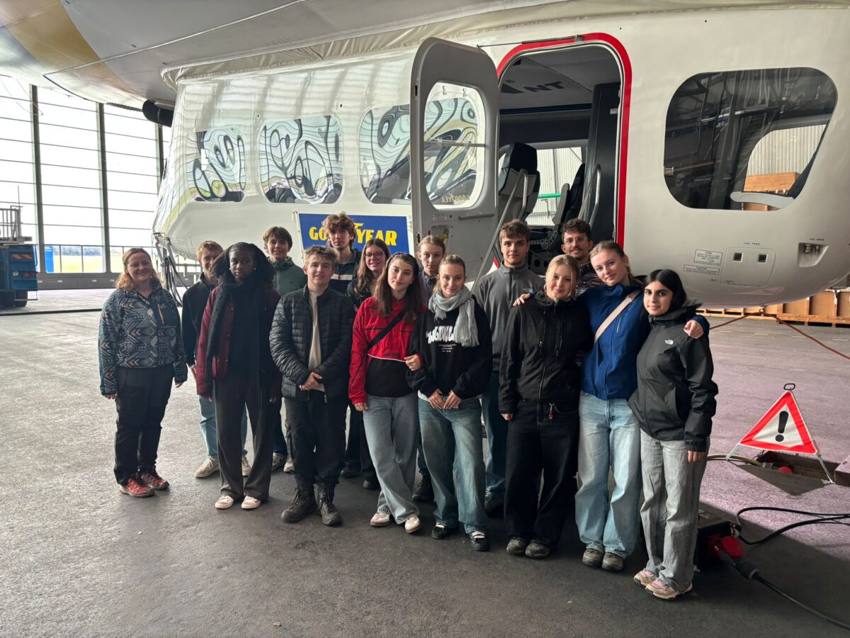 Eine Gruppe junger Menschen lächelt vor einem Luftschiff mit hellem Rumpf und Glasscheiben