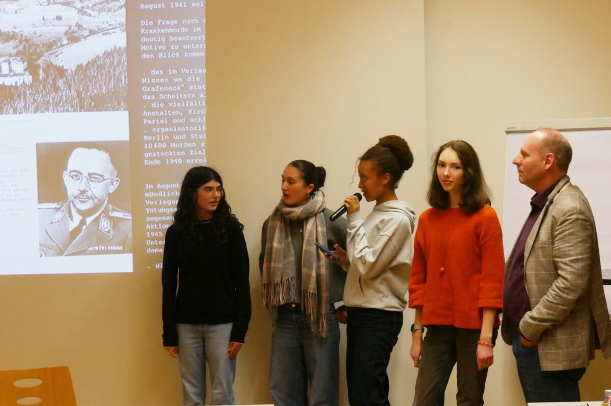 Fünf Personen stehen nebeneinander vor einer Präsentation, eine Person spricht ins Mikrofon