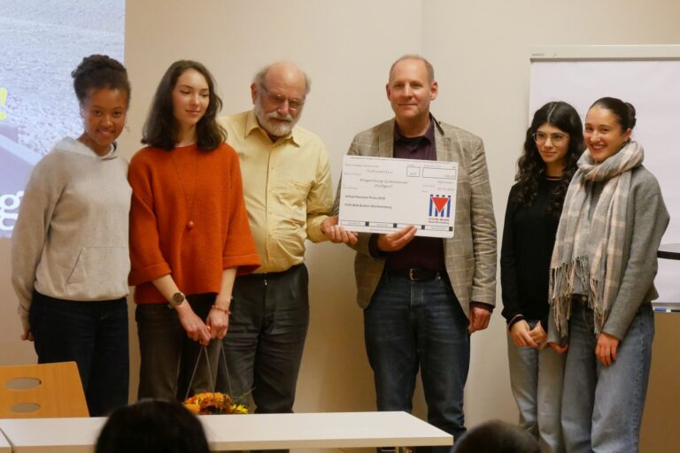 Sechs Personen stehen nebeneinander, eine Person hält ein großes weißes Schild und alle lächeln freundlich