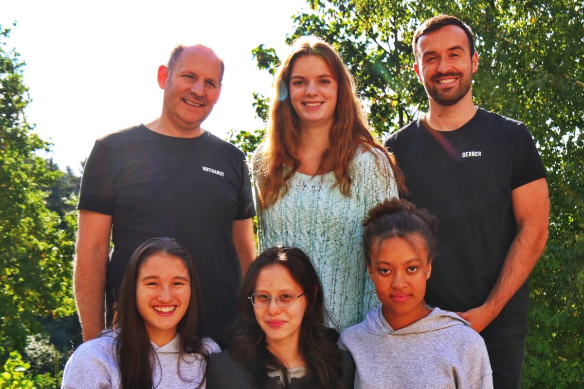 Drei Frauen und drei Männer lächeln freundlich im Sonnenlicht vor grünen Bäumen