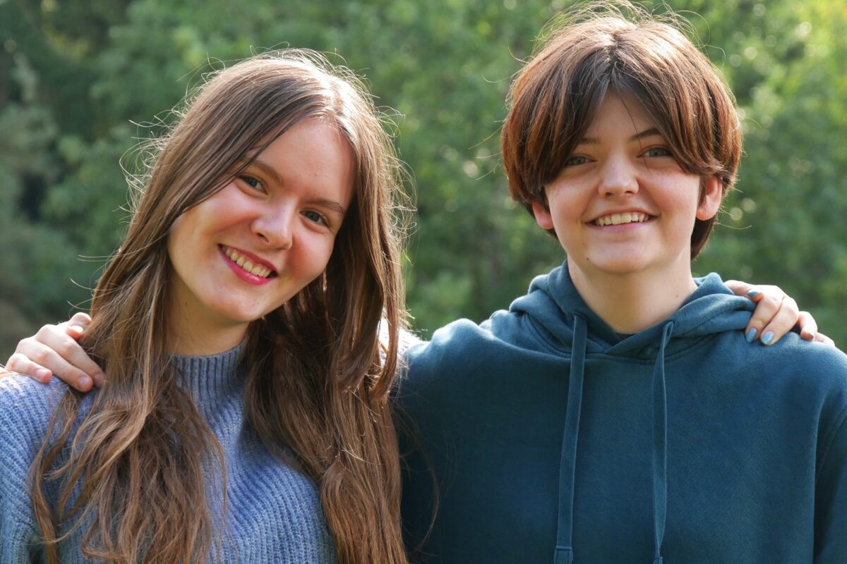 Zwei junge Frauen lächeln freundlich in die Kamera und stehen eng beisammen im Grünen