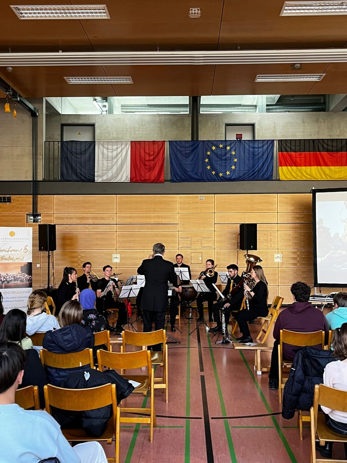 Ein Orchester spielt vor Publikum in einer Turnhalle mit Flaggen an der Wand