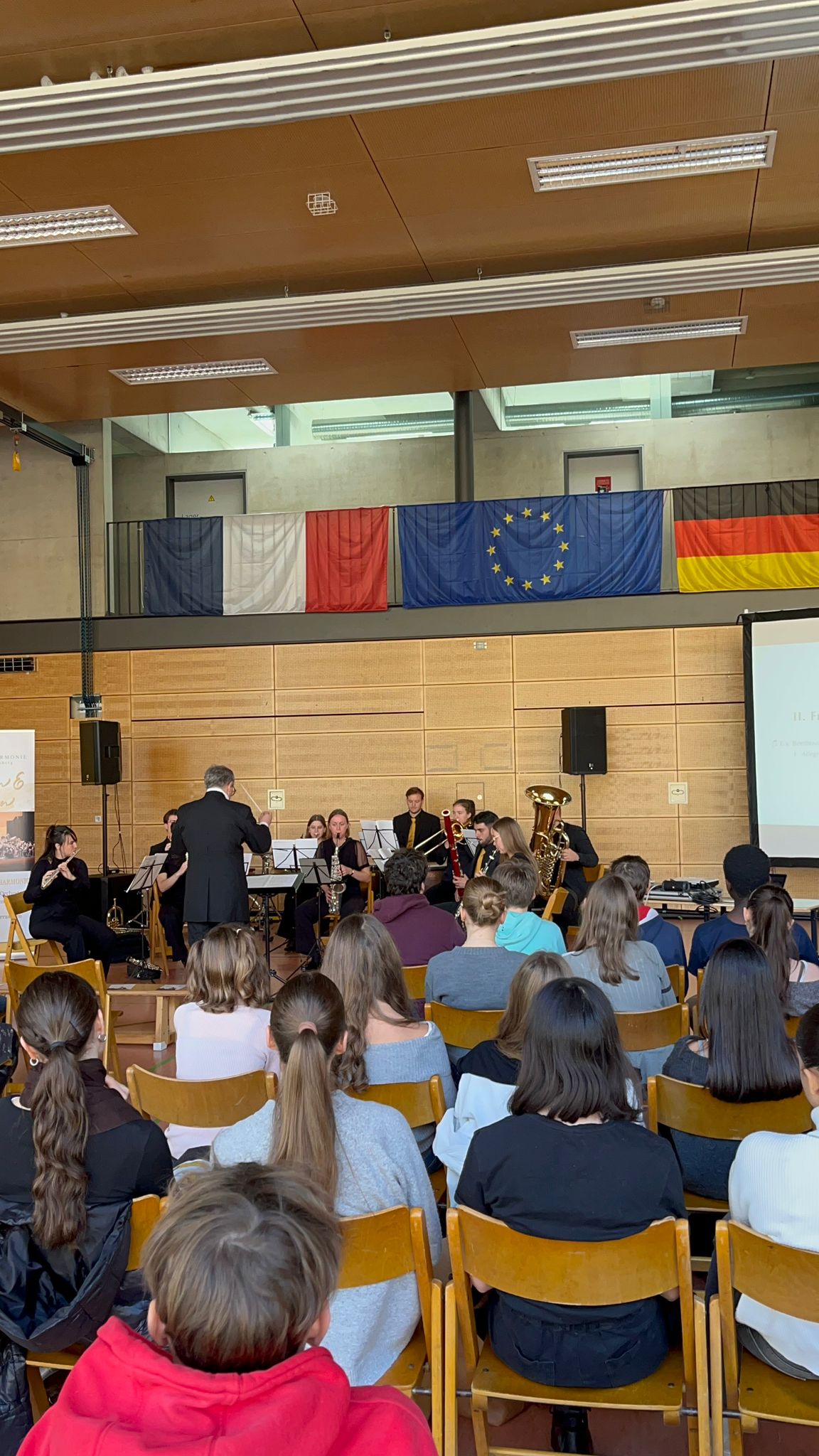 Eine Blaskapelle spielt in einer Turnhalle vor aufmerksamem Publikum unter Flaggen