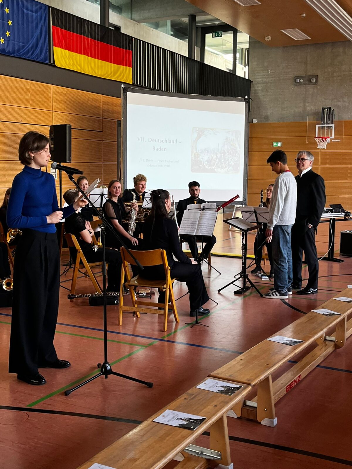 Eine junge Frau spricht freundlich ins Mikrofon vor einer sitzenden Schülerband in heller Turnhalle mit deutschen und EU-Flaggen im Hintergrund