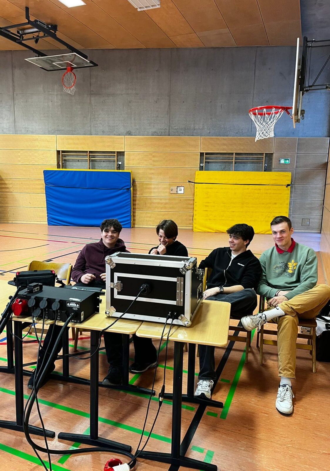 Vier junge Personen sitzen lächelnd an Tischen mit Technik in einer Turnhalle