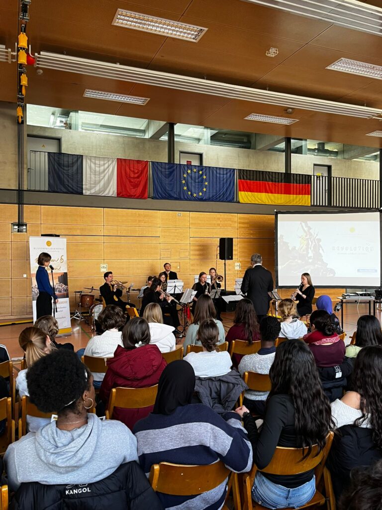 Ein gemischtes Publikum hört entspannt einem Orchester vor internationalen Flaggen zu