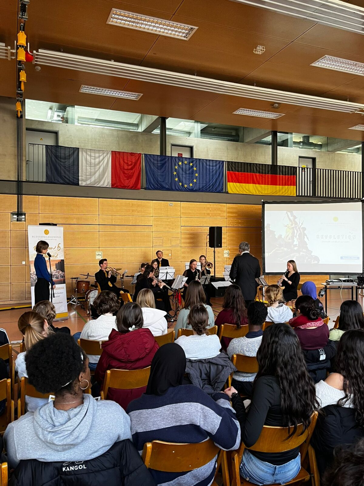 Ein gemischtes Publikum hört entspannt einem Orchester vor internationalen Flaggen zu