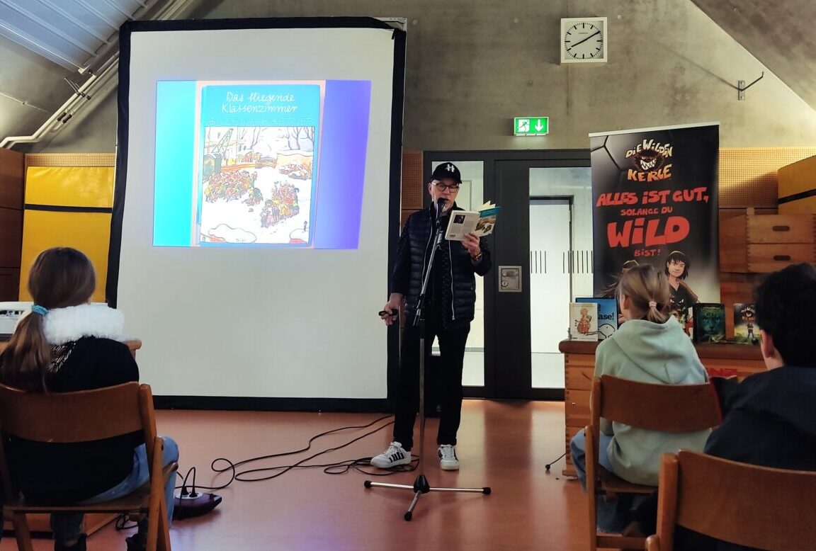 Eine Person mit schwarzer Kleidung liest freundlich aus einem Buch vor einer Leinwand vor