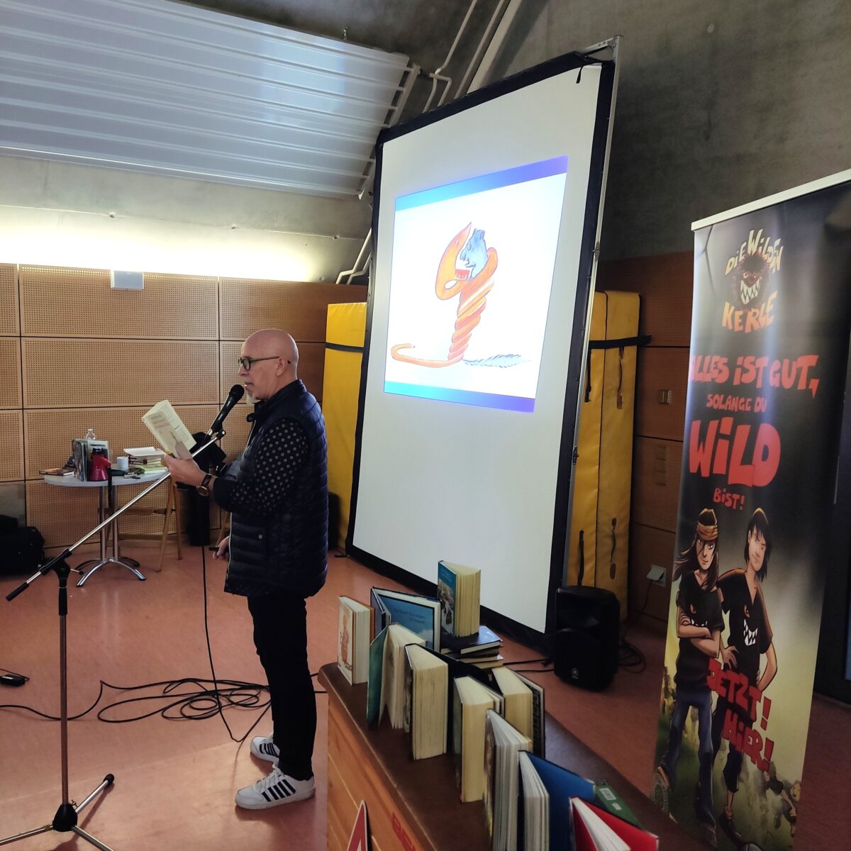 Ein Mann mit Brille liest lachend vor, daneben bunte Illustration einer Figur auf Leinwand