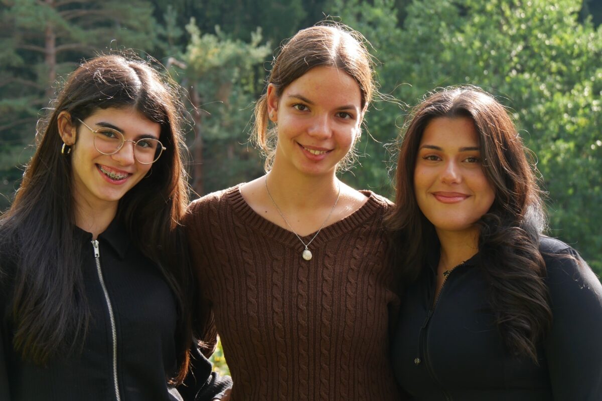 Drei junge Frauen lächeln herzlich, tragen dunkle Kleidung und stehen im Grünen